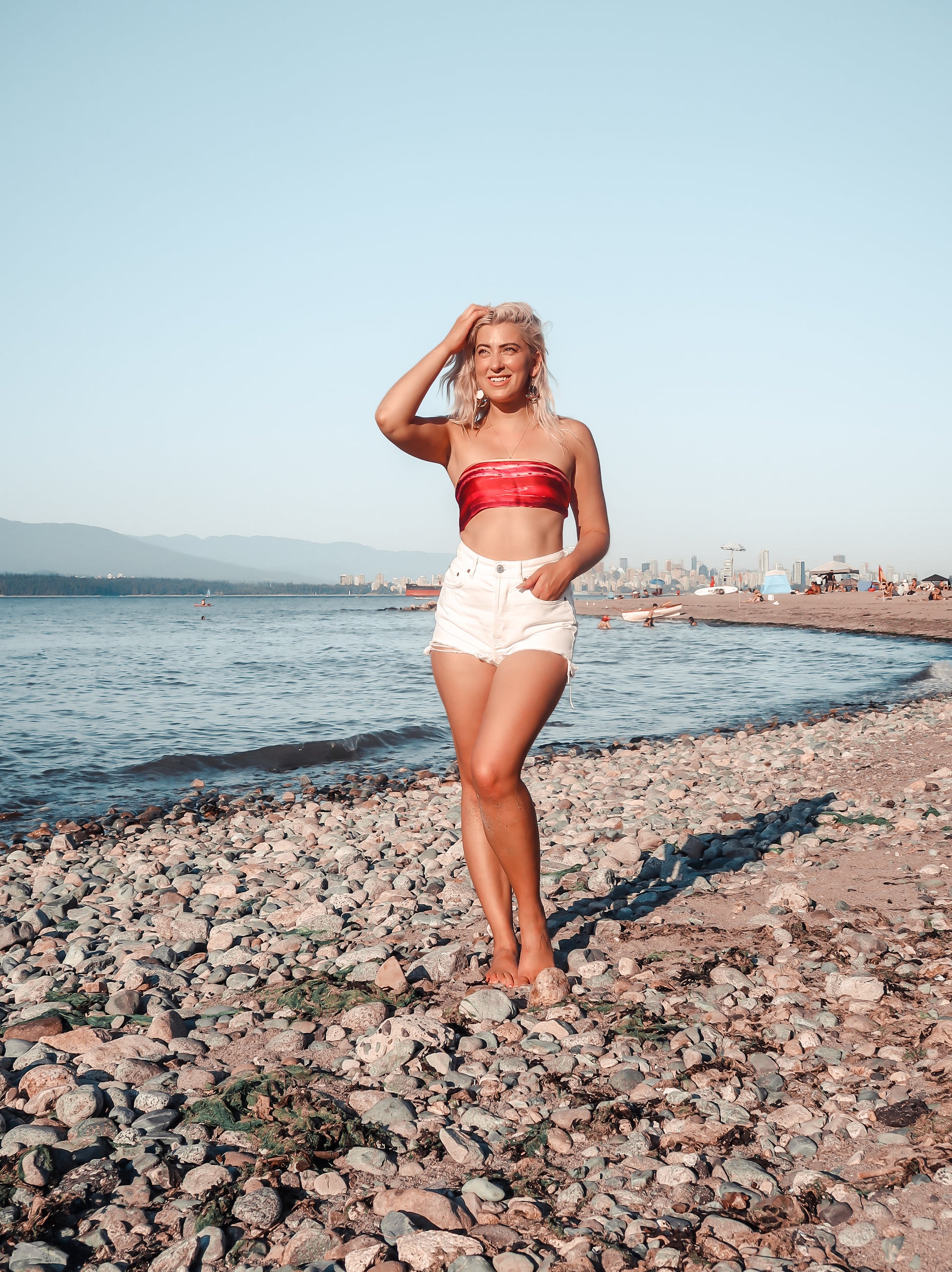 Woman standing on a rocky beach wearing a magenta bandeau top and white shorts.