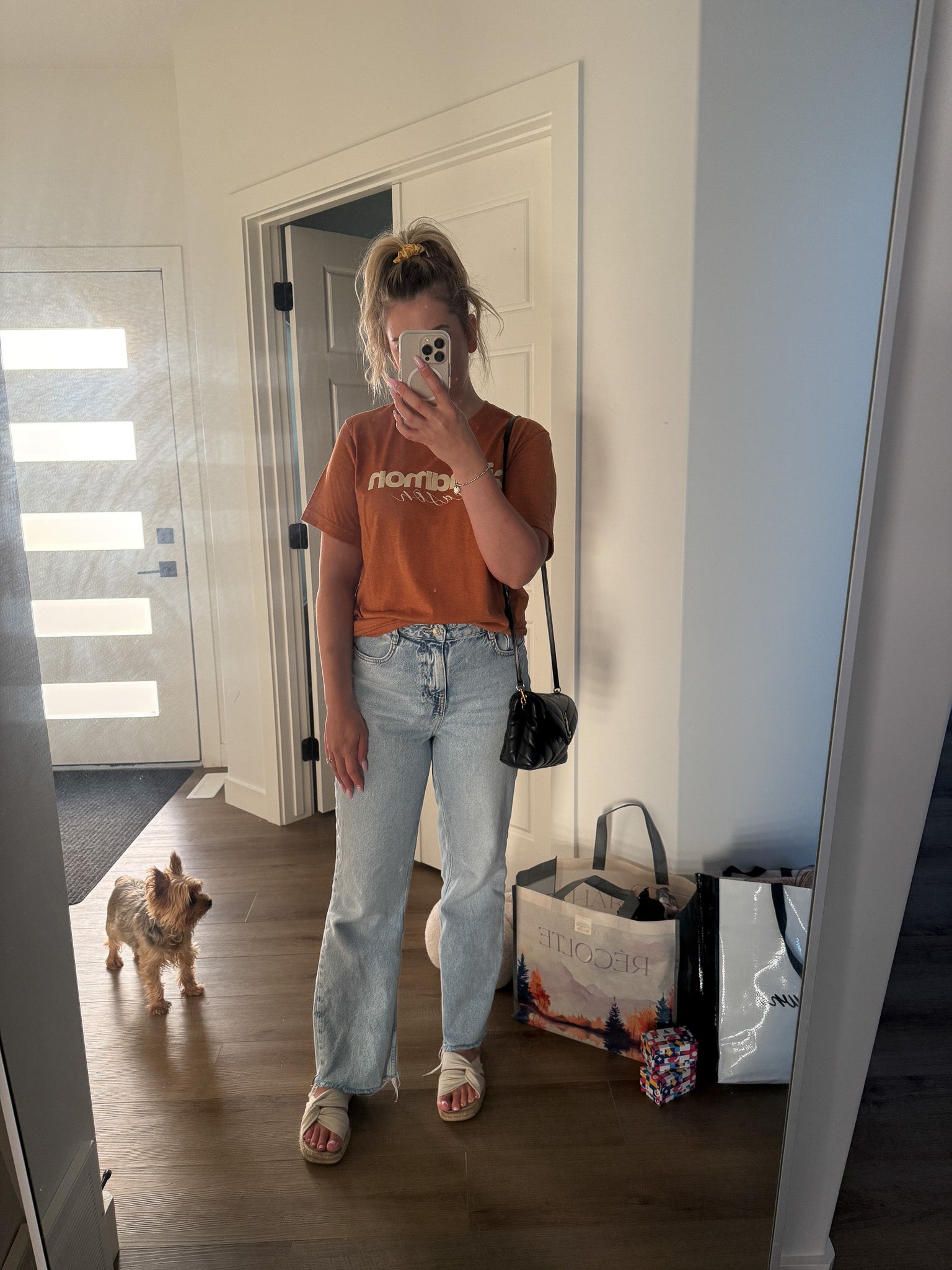 Person taking a mirror selfie wearing an orange t-shirt and light blue jeans, with a dog and bags in the background.