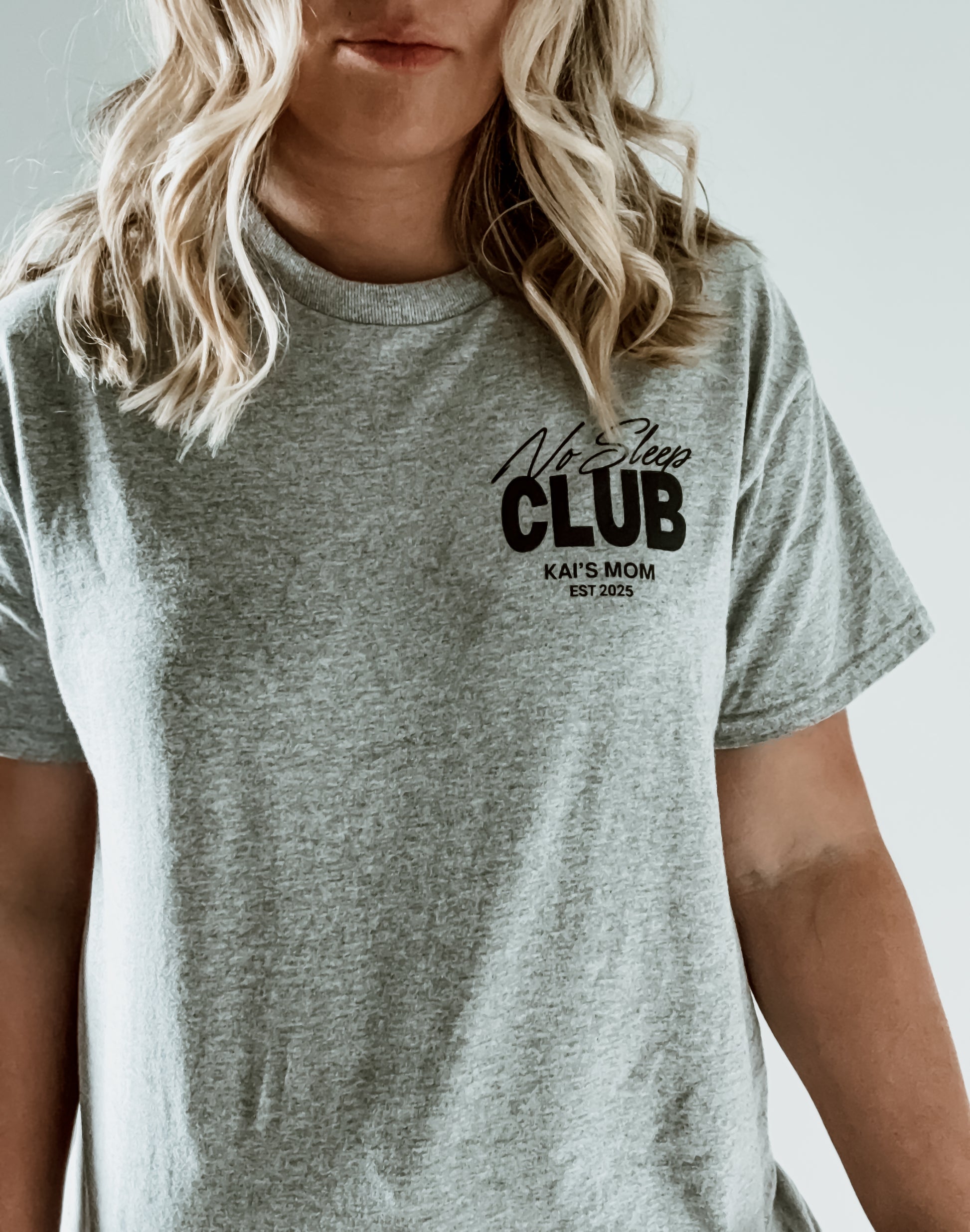 Person wearing a gray t-shirt with 'No Sleep Club Kai's Mom EST 2025' text.