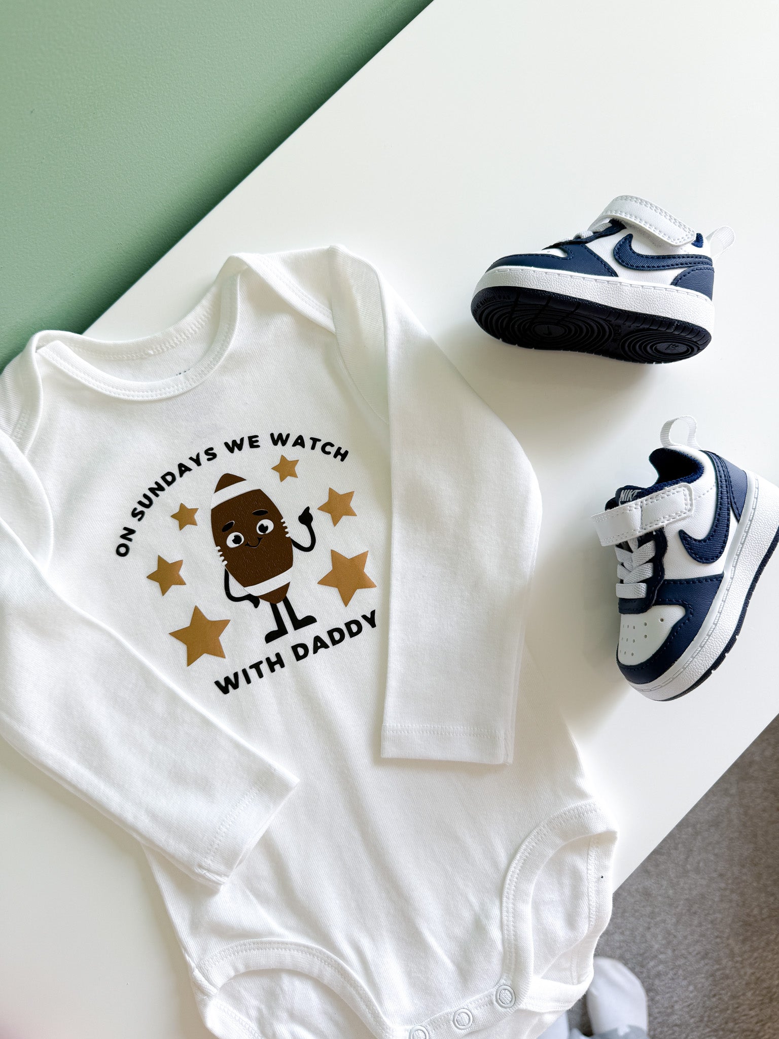 White baby onesie with a printed design and baby shoes on a light surface.