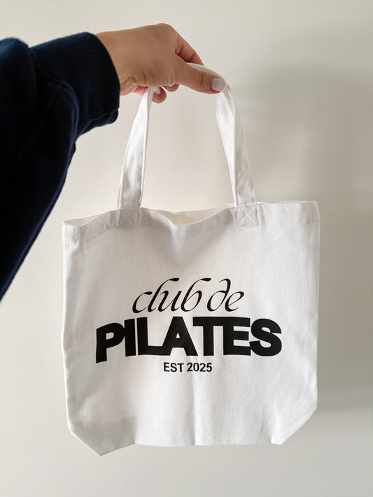 White tote bag with 'club de PILATES EST 2025' text held by a person against a neutral background
