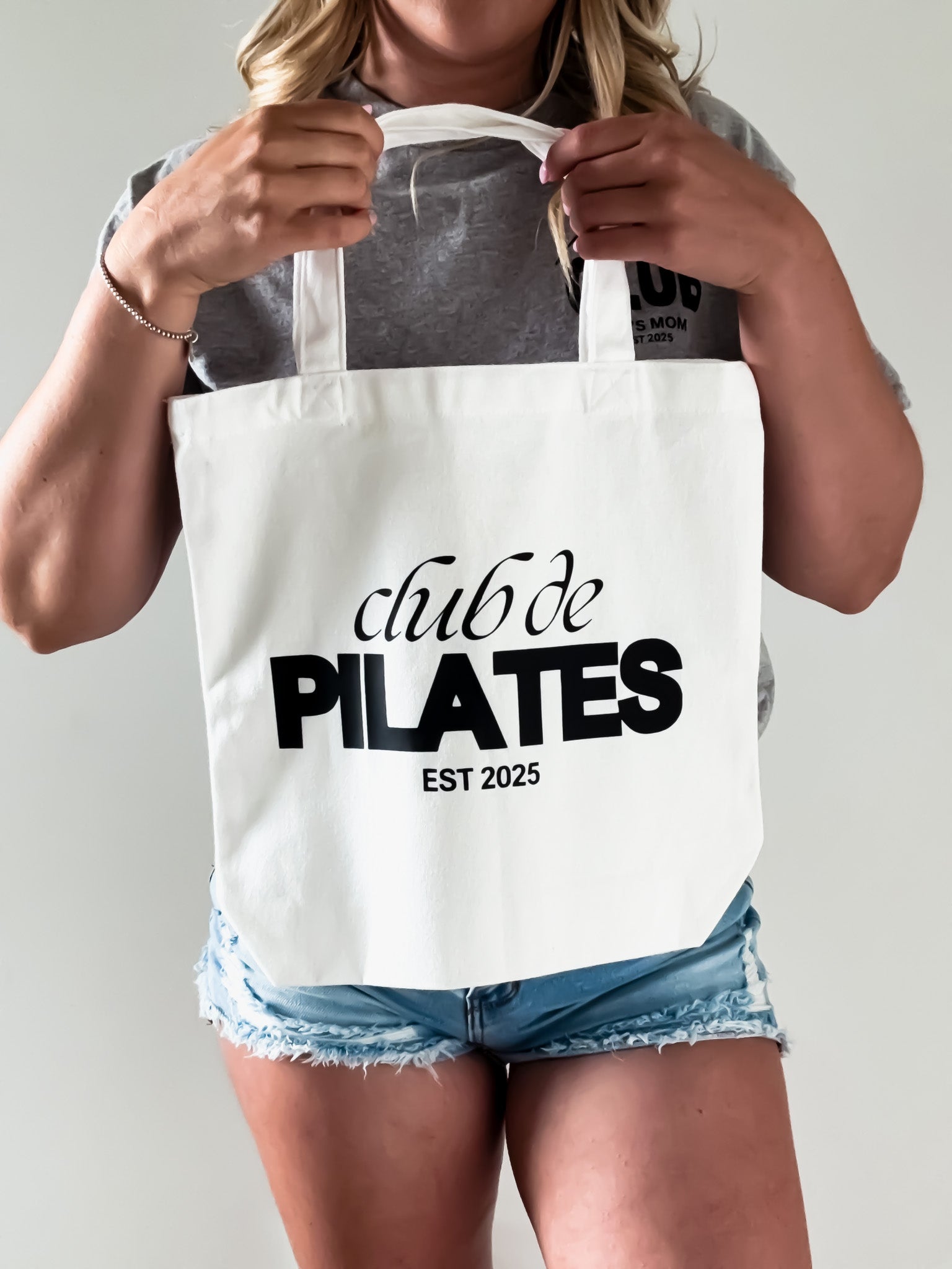 Person holding a tote bag with 'club de PILATES EST 2025' text on a plain background