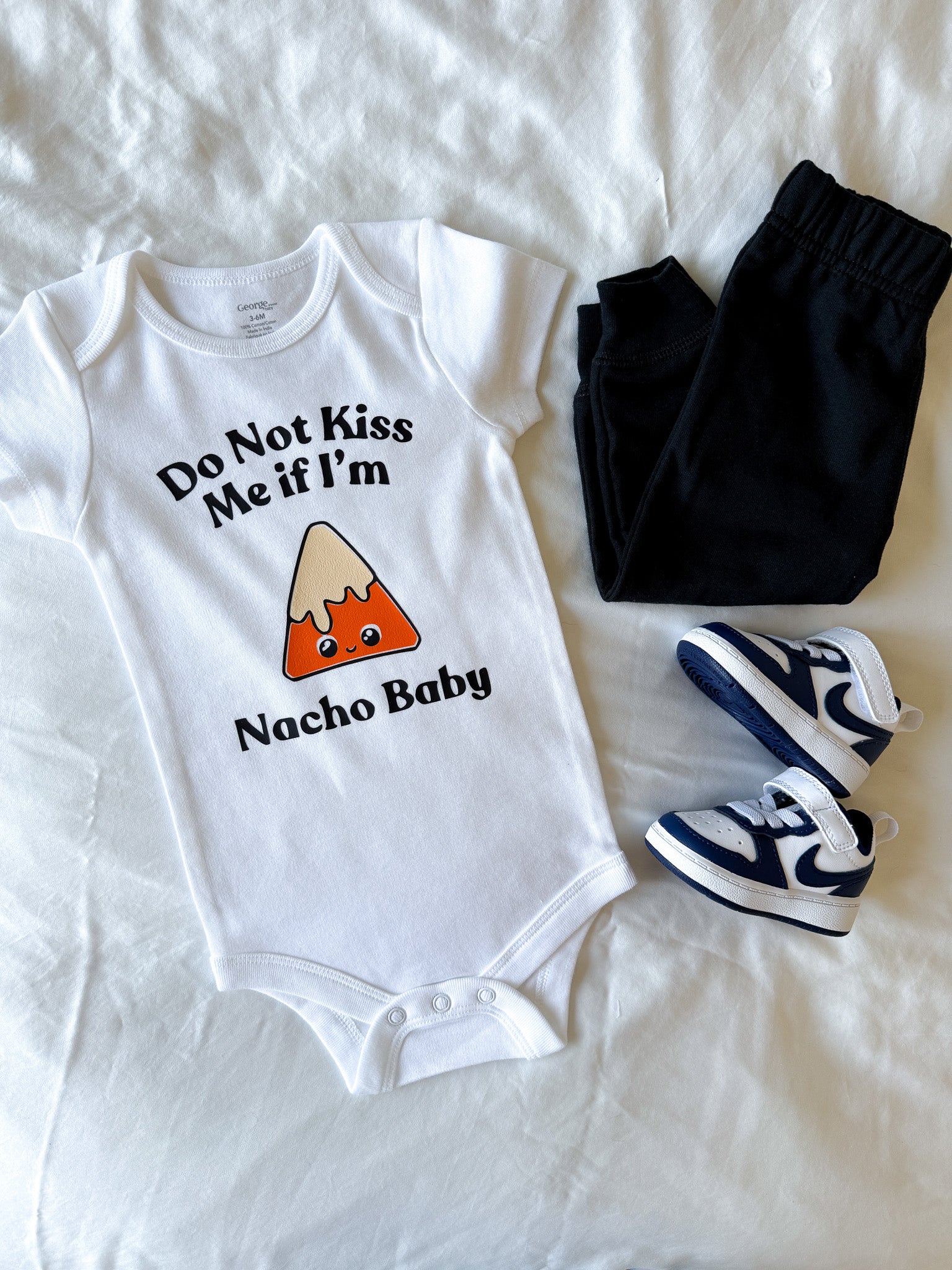 Baby onesie with 'Do Not Kiss Me if I'm Nacho Baby' text and nacho graphic, paired with black pants and blue sneakers on a white background.