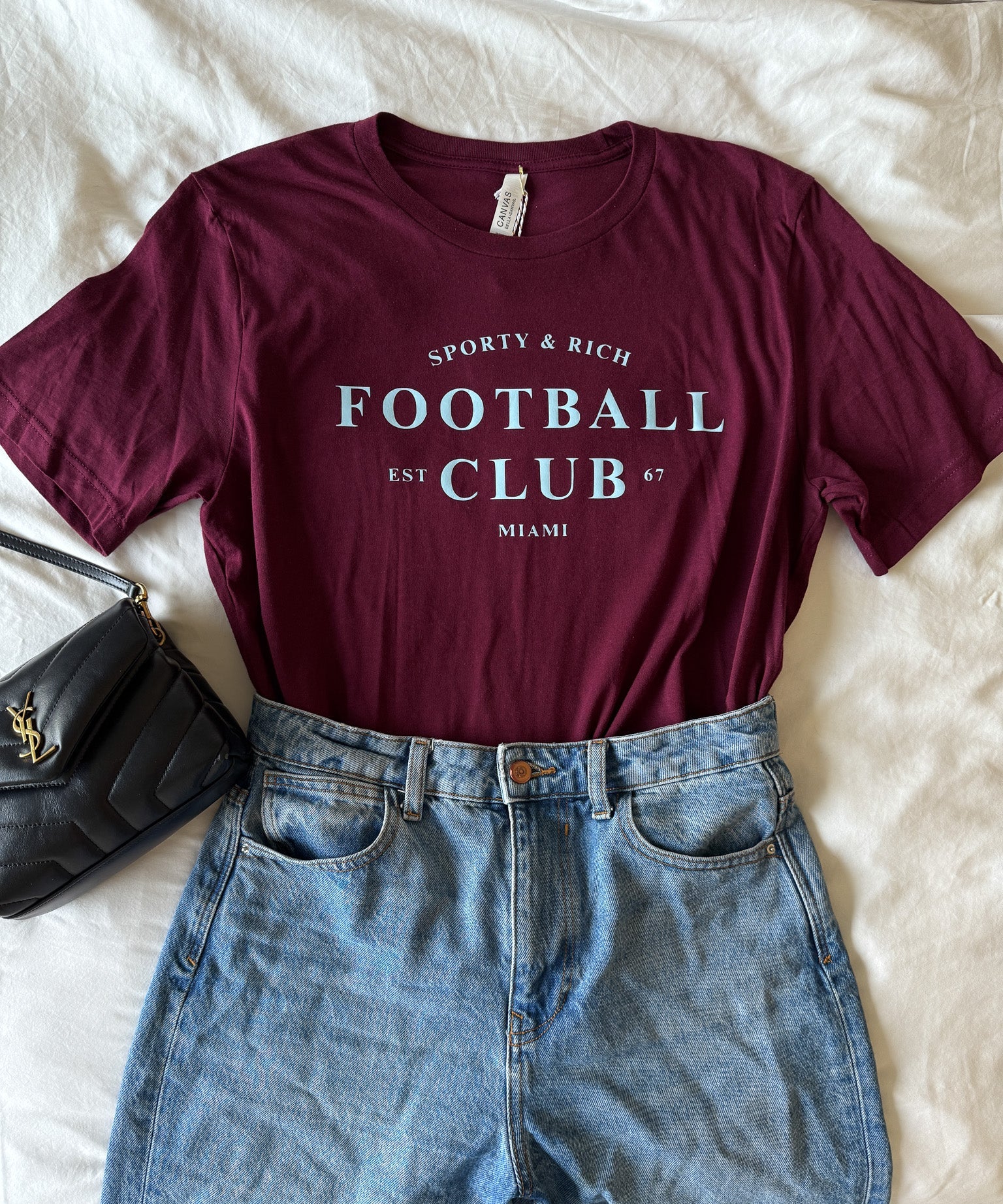 Maroon t-shirt with 'Football Club' text and blue jeans on a white surface