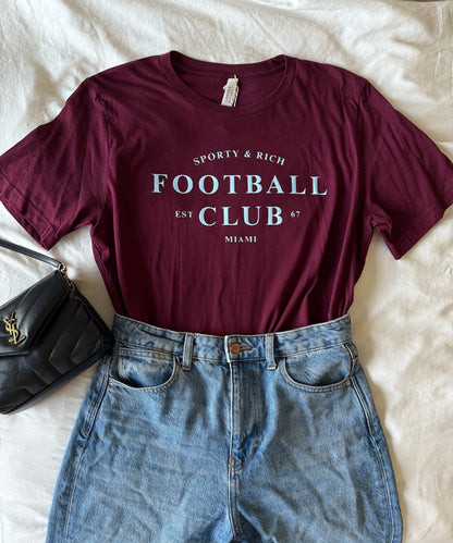 Maroon t-shirt with 'Football Club' text and blue jeans on a white surface