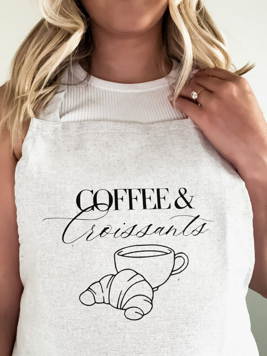 Grey apron with 'Coffee & Croissants' design on a person wearing it.