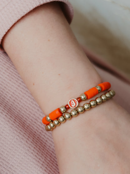 Close-up of a wrist wearing two bracelets, one orange and one gold, on a soft pink background.