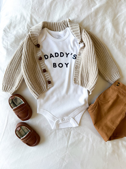 Baby outfit with white 'DADDY'S BOY' onesie, beige cardigan, brown pants, and shoes on a white background