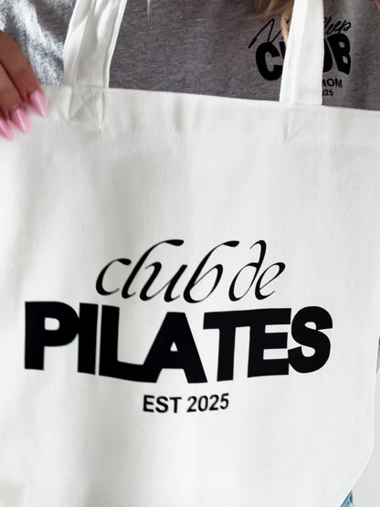 White tote bag with 'club de PILATES EST 2025' text held by a person wearing a gray shirt with similar text.
