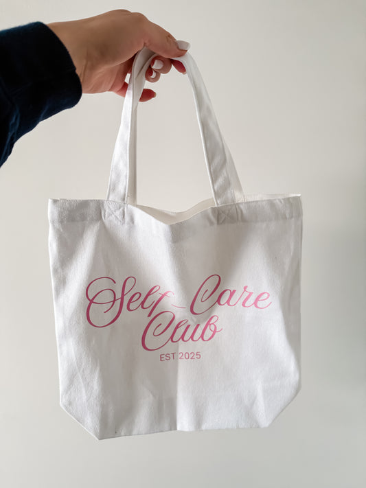 White tote bag with 'Self Care Club' text held by a hand against a plain background