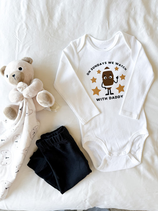 Baby onesie with printed design, black pants, and teddy bear on a white blanket