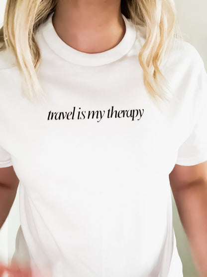 White t-shirt with 'travel is my therapy' text worn by a person.