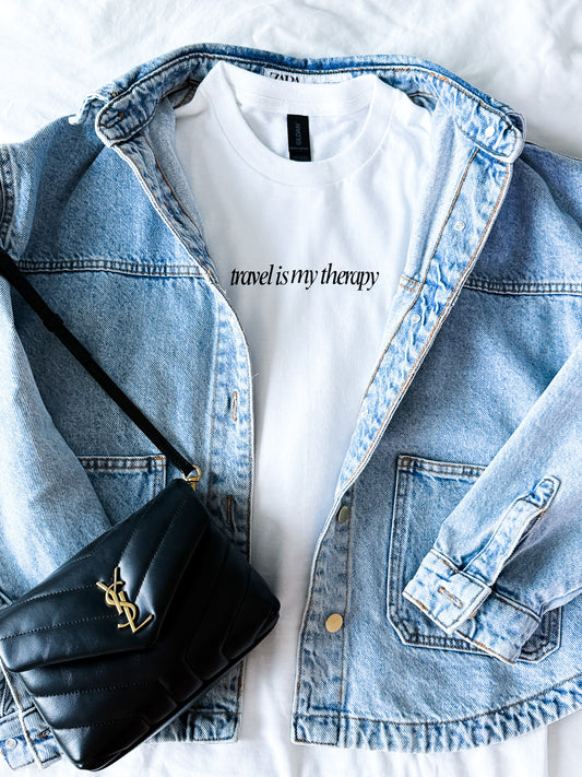 White t-shirt with 'travel is my therapy' text, denim jacket, and black handbag on a white background