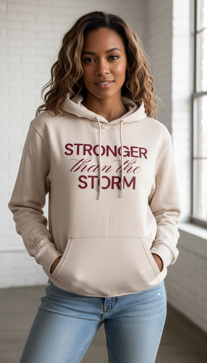 Woman wearing a beige hoodie with 'Stronger than the storm' text in a room with large windows.